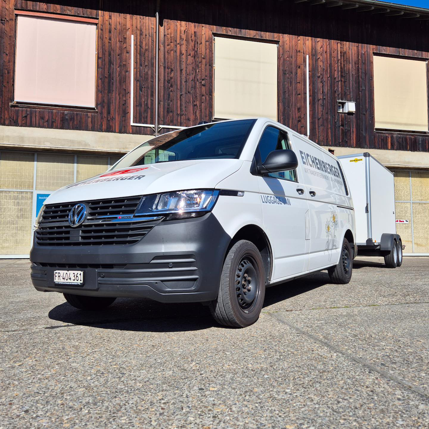 Weißer Volkswagen Transporter mit Anhänger vor Holzgebäude